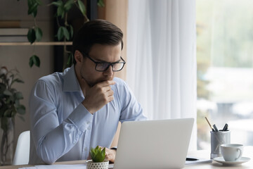 Serious focused businessman in eyeglasses working on laptop sit at workplace desk, read e-mail, feels concerned analyze received information. Challenge, new app or software learning, brainwork concept