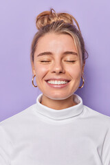 Sticker - Vertical shot of pleased attractive woman smiles toothily keeps eyes closed imagines something wears white jumper isolated over purple background. People happy emotions and feelings concept.