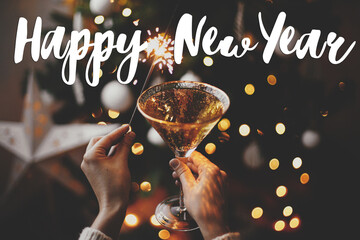 Wall Mural - Happy New Year text on hands holding burning sparkler and champagne glass on background of christmas tree lights. Season's greetings card. Handwritten sign. Happy holidays