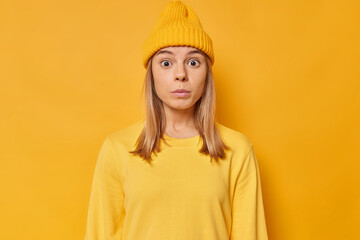 Wall Mural - Shocked impressed young European woman stares bugged eyes feels fascinated hears something astonishing wears hat and casual jumper isolated over vivid yellow background. Human reactions concept