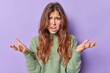 Irritated displeased young European woman keeps palms sideways looks clueless and dissatisfied has long dark hair exclaims from unhappiness wears casual jumper isolated over purple background.