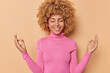 © wayhome.studio  - Horizontal shot of happy young woman with curly hair keeps eyes closed smiles broadly meditatees and makes zen gesture tries to reach nirvana smiles pleasantly wears pink jumper poses indoor