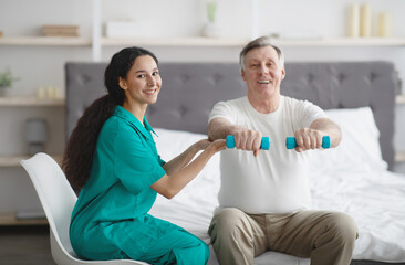 Wall Mural - Female physiotherapist helping positive mature man to do exercises with dumbbells on bed at retirement home