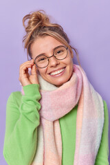 Wall Mural - Indoor shot of lovely young fair haired woman tilts head keeps hand on rim of spectacles smiles broadly looks joyfully at camera hears good news wears long sleeved jumper and warm scarf around neck