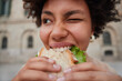 © WHstudio Leushin N - Close up shot of positive curly haired glad young woman winks eye holds appetizing sandwich with hands bites delicious snack returns from fast food restaurnat poses outdoors. Cheat meal concept.