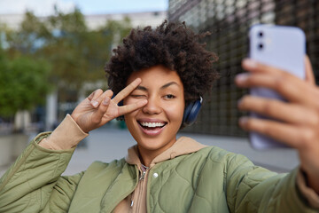 Wall Mural - beautiful smiling woman makes peace gesture over eye has carefree expression takes selfie via smartp