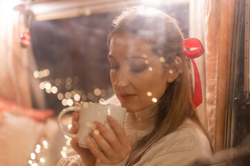 blurry portrait of candid woman face through the window glass holding in hands and enjoyment cup of cocoa drink and relaxing on happy new year and merry christmas eve. bokeh of glowing lights garland