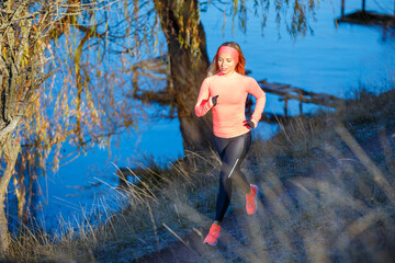 Wall Mural - Young sporty woman in orange longsleeve running along blue frosty lake in the morning.