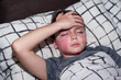 a boy of European appearance with red spots on his face is lying in bed with his hand on his forehead
