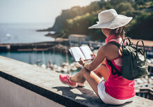 A Woman Sitting On Her Back Looking At A Travel Diary. She Is By The Sea. She Is Wearing A White Cap And Shorts. Concept Travel And Free Time.