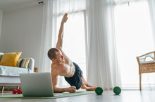 Young Muscular Topless Body Man Making Side Plank Exercise Following Online A Fitness Trainer Using A Laptop. Home Workout In The Living Room. Healthy Lifestyle And Modern Online Technology Concept.