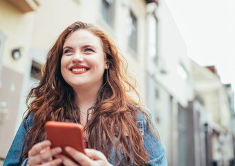 Wall Mural - Smiling red curly long hair caucasian teen girl walking on the street and browsing the internet using the modern smartphone. Modern people with technology devices concept image.