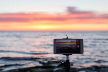 Sunrise Over The Sea Coast In A Colorful Orange Sky. Smartphone Camera On A Tripod In The Foreground.
