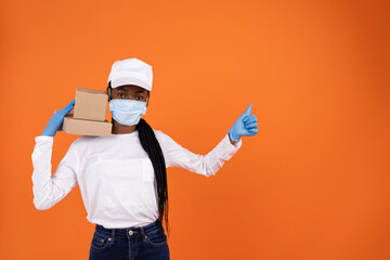 Wall Mural - African-American delivery lady in a virus mask and blue gloves holding boxes on her shoulder and showing her thumbs up on orange background with copy space