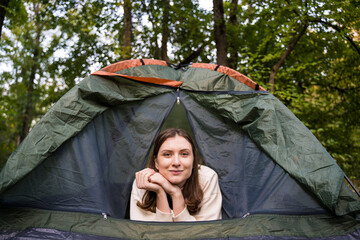 Portrait of a tourist woman peeking out of a green tent in the woods in nature. Adventure, journey into the forest