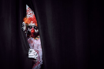 Terrifying clown hiding behind black curtains, space for text. Halloween party costume