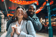 Man Covering Eyes Of Girlfriend At Illuminated Christmas Market