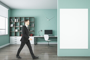 Poster - Businessman walking in modern blue office interior with empty mock up poster on wall, bookcase, window with city view, workplace with computer, other items and wooden flooring.