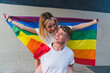 © Westend61 - Happy woman piggybacking girlfriend with rainbow flag in front of wall