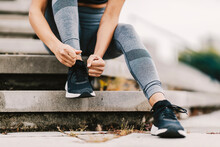 Close Up Of Female Runner Sitting On The Stairs Outdoors And Tying A Shoelace On Her Sneaker. A Runner Getting Ready For Running