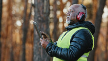 Elderly African American Forestry Engineer In Noiseisolating Headphones In Forest Old Mature Foreman Forester Thinks Over Work Plan For Cutting Trees By Entering Data Into Tablet Evaluating Situation