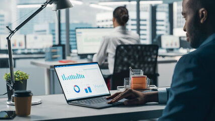 Poster - Successful Black Businessman Sitting at Desk Working on Laptop Computer in Big City Office. Hard Working Top Manager Doing Research for e-Commerce Project with Graps, Charts and Tables. Over Shoulder