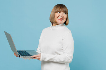 Wall Mural - Elderly copywriter woman 50s wear white knitted sweater hold use work on laptop pc computer look aside on workspace isolated on plain blue color background studio portrait. People lifestyle concept