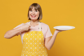 Elderly fun housekeeper housewife woman 50s in orange apron point finger on empty plate with copy space workspace area isolated plain on yellow background studio. People household lifestyle concept.