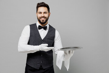 Young fun barista male waiter butler man wear white shirt vest elegant uniform work at cafe show hold in hand carrying metal tray isolated on plain grey background studio. Restaurant employee concept.