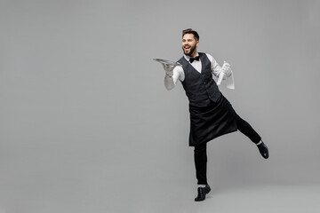 Wall Mural - Full size young smiling barista male waiter butler man wear white shirt vest elegant uniform work at cafe carrying walk give metal tray isolated on plain grey background Restaurant employee concept.