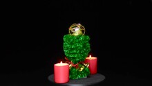 Between Two Red Burning Candles, A Golden Christmas Ball Rotates On A Green Decorated Stand