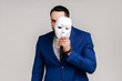 © khosrork - Bearded man holding white mask, covering face, standing with serious expression, multiple personality disorder, wearing official style suit. Indoor studio shot isolated on gray background.