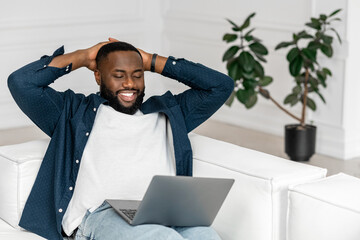 Serene African-American guy in casual shirt over t-shirt taking a break, lying on the sofa with a laptop on his lap, holding hands behind head and rest, spending leisure at home alone