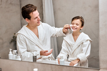 Man in bathroom putting shaving cream on himself and child son, in bathrobes. man and little boy with shaving foam on faces looking in bathroom mirror and laughing. Father and son have fun