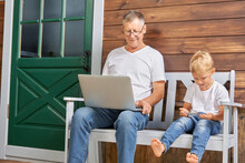 Grandfather With A Laptop And A Grandson With A Smartphone Are Sitting On Benches Near A Wooden House. Concept Of Using Modern Gadgets: Laptop, Smartphone And Internet In The Village