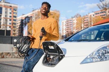 Wall Mural - Electric cars, EV concept, eco friendly fuel. Portrait of young smiling black man, recharging his modern luxury electric car