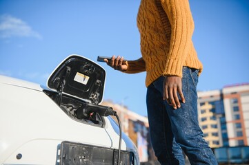 Wall Mural - Close up of african american man connecting charging cable to electric car. Young male standing near his modern auto with leather suitcase in hand.