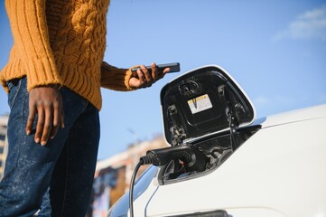 Close up of african american man connecting charging cable to electric car. Young male standing near his modern auto with leather suitcase in hand.