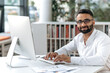 © Kateryna - Smart successful handsome Indian businessman, manager, office worker with beard and glasses. Business owner sitting at table in modern office, using computer, looking at camera, smiling