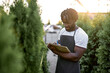 © Roman - Focused black male writing notes at plants nursery, wearing uniform. man checking quality of plants and take notes on paper in garden greenhouse, Agricultural fresh organic concepts. at sunny day
