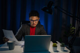Fototapeta  - Portrait of tired focused young man in glasses working remotely late at night at home office with computer, looking at laptop, thinking about deadline. Overwork