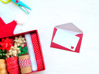 White card with a heart hole punch and a red envelope with  red paper box with christmas decorations, scissors, santa hats on white wooden background. Top view, with copy space, New year concept..