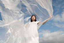 Young Asian Woman In White Dress Holding Plastic Sheet In Wind Under Blue Sky