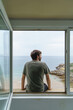 © beavera - Back view of unrecognizable man sit on a window frame looking at the sea. Horizontal view of man isolated at home with a view from the window of the blue ocean on the background. People outdoors