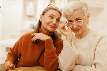 Wall Mural - People, aging, maturity and lifestyle concept. Beautiful short haired retired woman in stylish white glasses sitting at cafe table with her best friend, waiting for coffee, discussing future plans