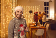 Portrait of happy, smiling young man wearing traditional hat and ugly Christmas sweater with snowman holding wine glass standing against background of decorated interior at festive party with friends
