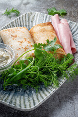 Poster - Pancakes filled with ham in a plate with parsley and dill greens and sour cream on a gray stone table