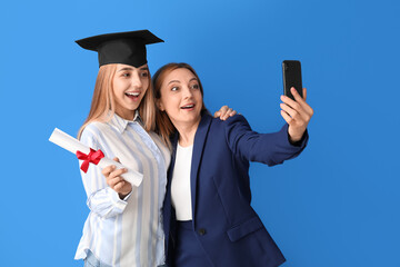 Wall Mural - Happy female graduation student with her mother taking selfie on color background