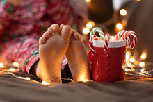 Close Up Child Bare Feet Kid In Red Christmas Pajamas Hold Mug Cup Striped Green Red Candy Christmas Canes Bokeh Lights Background. New Year