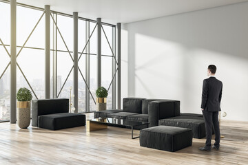 Poster - Thoughtful businessman standing in contemporary living room or business hall interior with sunlight, window with panoramic city view, furniture and mock up place on concrete wall, wooden flooring.